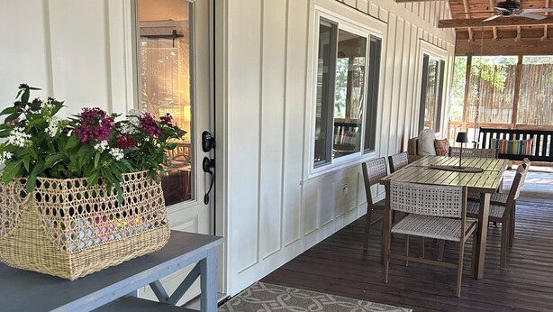 Une véranda abritée avec une table et des chaises, un panier en osier rempli de fleurs et une porte donnant sur l’intérieur.