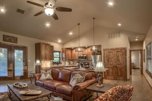 A spacious living room with a brown leather sofa, a coffee table, and a dining area with wooden cabinets and a kitchen in the background. - Lone Star Cabin No. 5 (Fredericksburg)