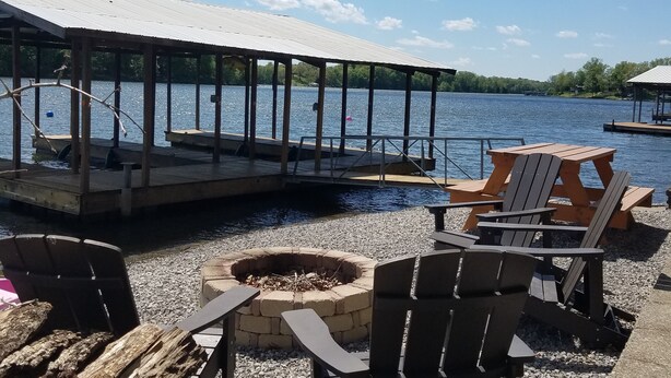 Una terraza junto al lago con sillas Adirondack, un pozo de fuego y un pabellón cubierto.