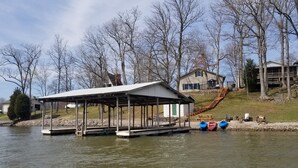 Un muelle a orillas del lago con una zona de estar cubierta, una casa con una escalera que la conduce y una fila de árboles al fondo.