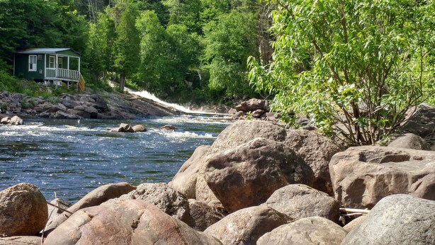 Un chalet au bord de la rivière, doté d’une véranda et entouré de gros rochers et d’un feuillage verdoyant et dense.