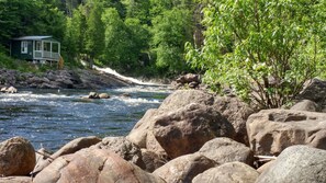 Un chalet au bord de la rivière, doté d’une véranda et entouré de gros rochers et d’un feuillage verdoyant et dense.