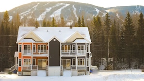 Une maison à deux étages, avec une partie centrale de couleur sombre et des murs extérieurs orange et blancs, se dressant sur fond de montagnes enneigées.