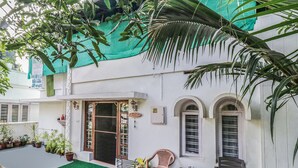 Un edificio blanco con techo verde, una puerta y ventanas. Hay plantas en macetas afuera y una silla adentro.