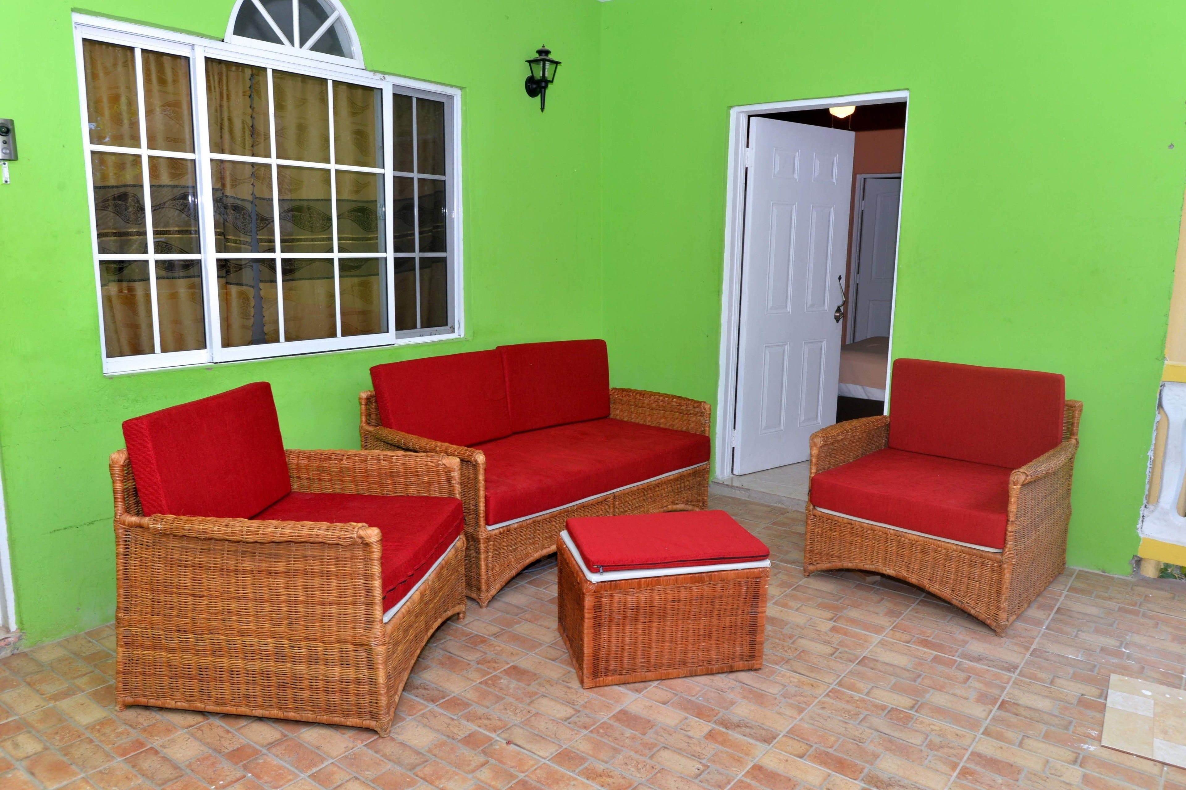 A room with two wicker sofas with red cushions, a small ottoman, a window with curtains, and a door leading to another room.