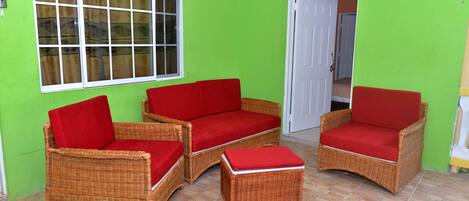 A room with two wicker sofas with red cushions, a small ottoman, a window with curtains, and a door leading to another room.