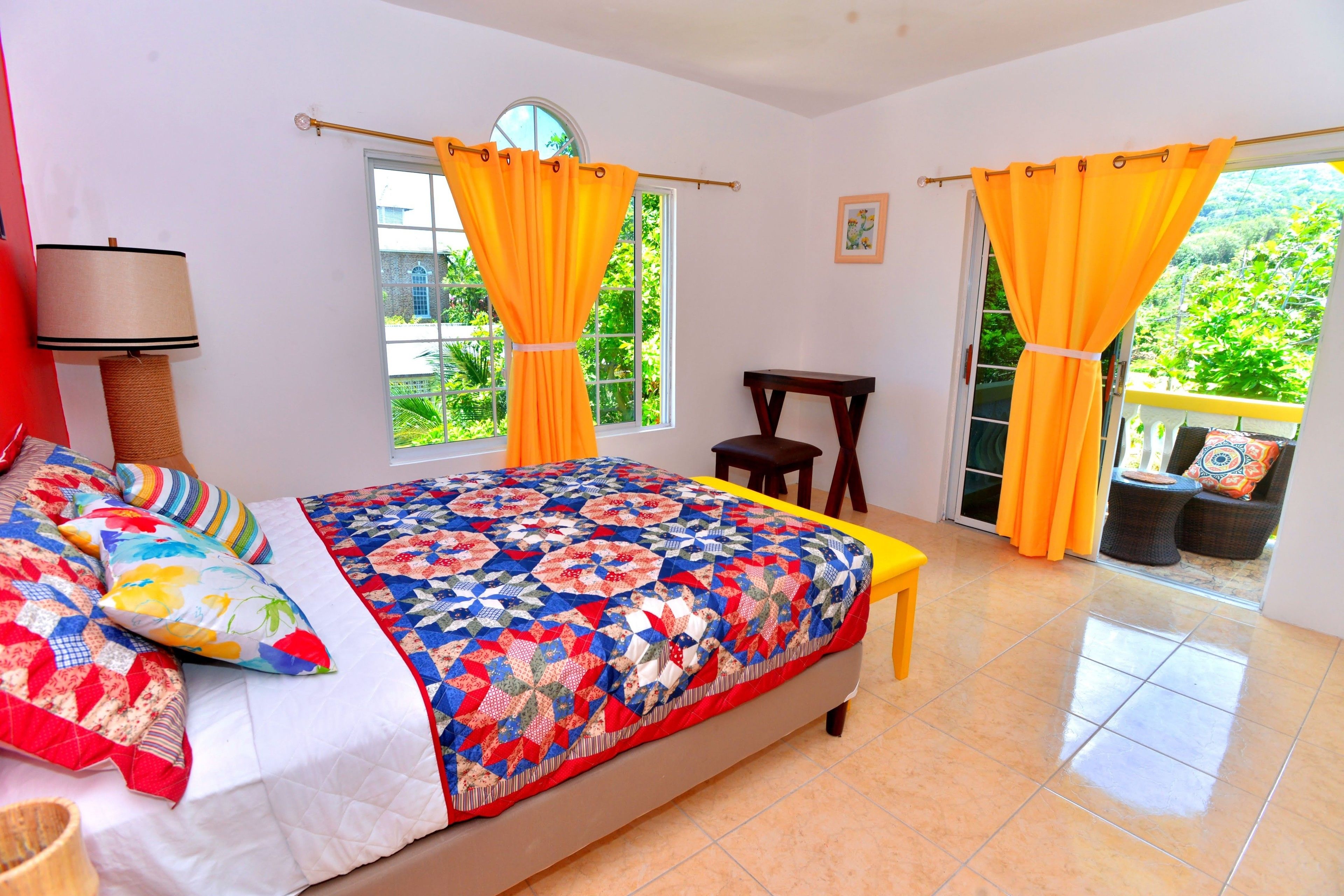 A bedroom with a large bed, two windows with curtains, a bedside table, and a view of greenery outside.