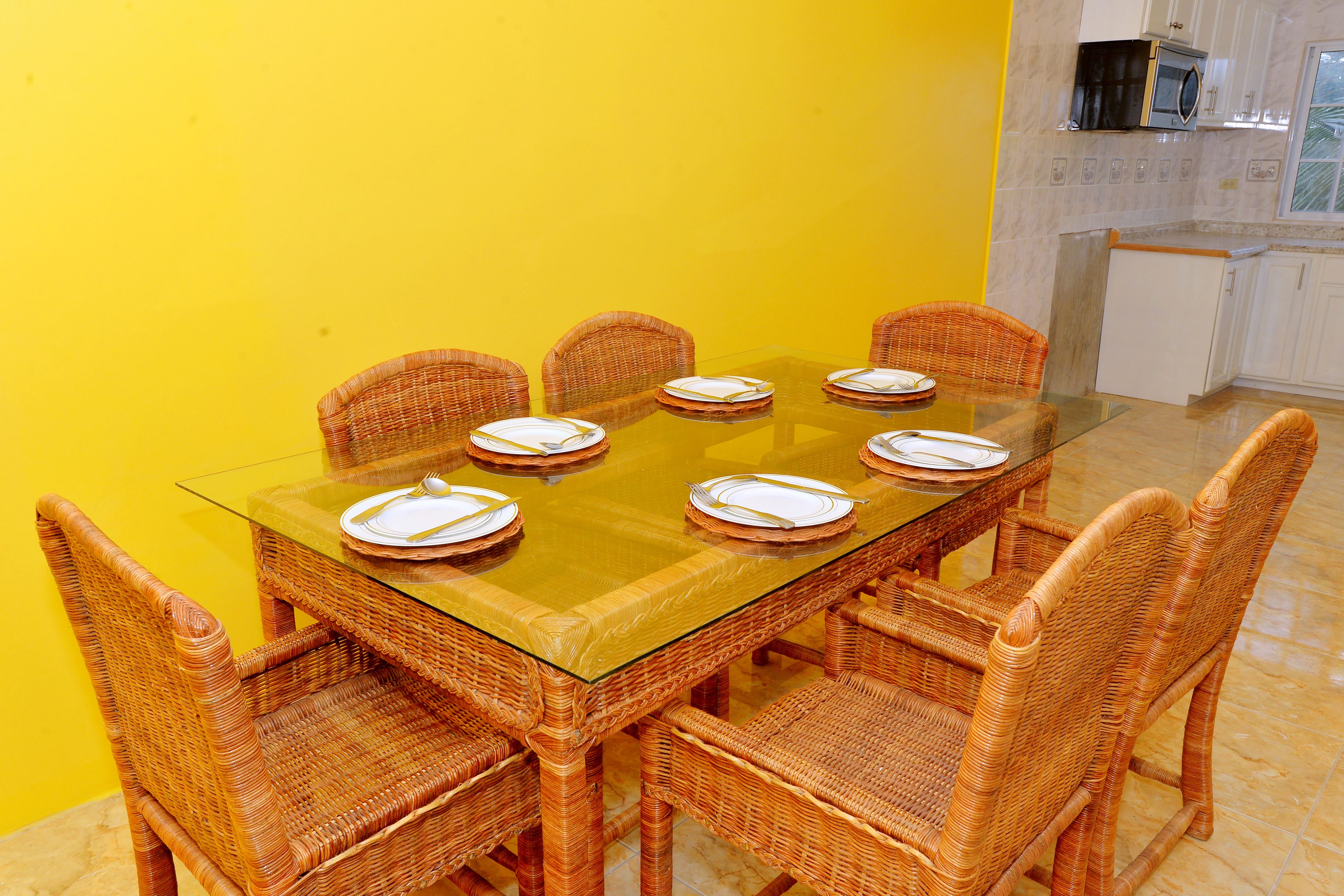 A dining area with a glass-top table, wicker chairs, and a yellow wall.