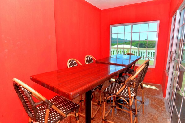 A dining area with a long wooden table and wicker chairs, a large window with a view of greenery, and a red wall.