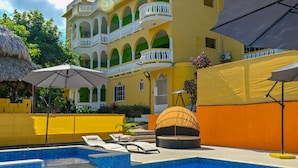 A swimming pool with lounge chairs and umbrellas, surrounded by a yellow building with balconies and a thatched roof structure. - Takuma Boutique Hotel Rooms & Suites Jamaica  (Montego Bay)