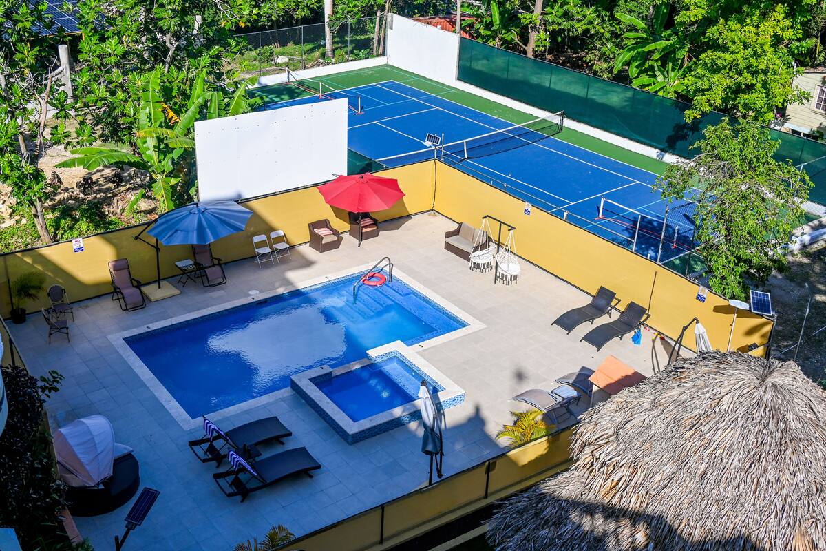 Ein Außenpoolbereich mit blauem Pool, Liegestühlen, einem roten Sonnenschirm und einer Struktur mit einem Reetdach.