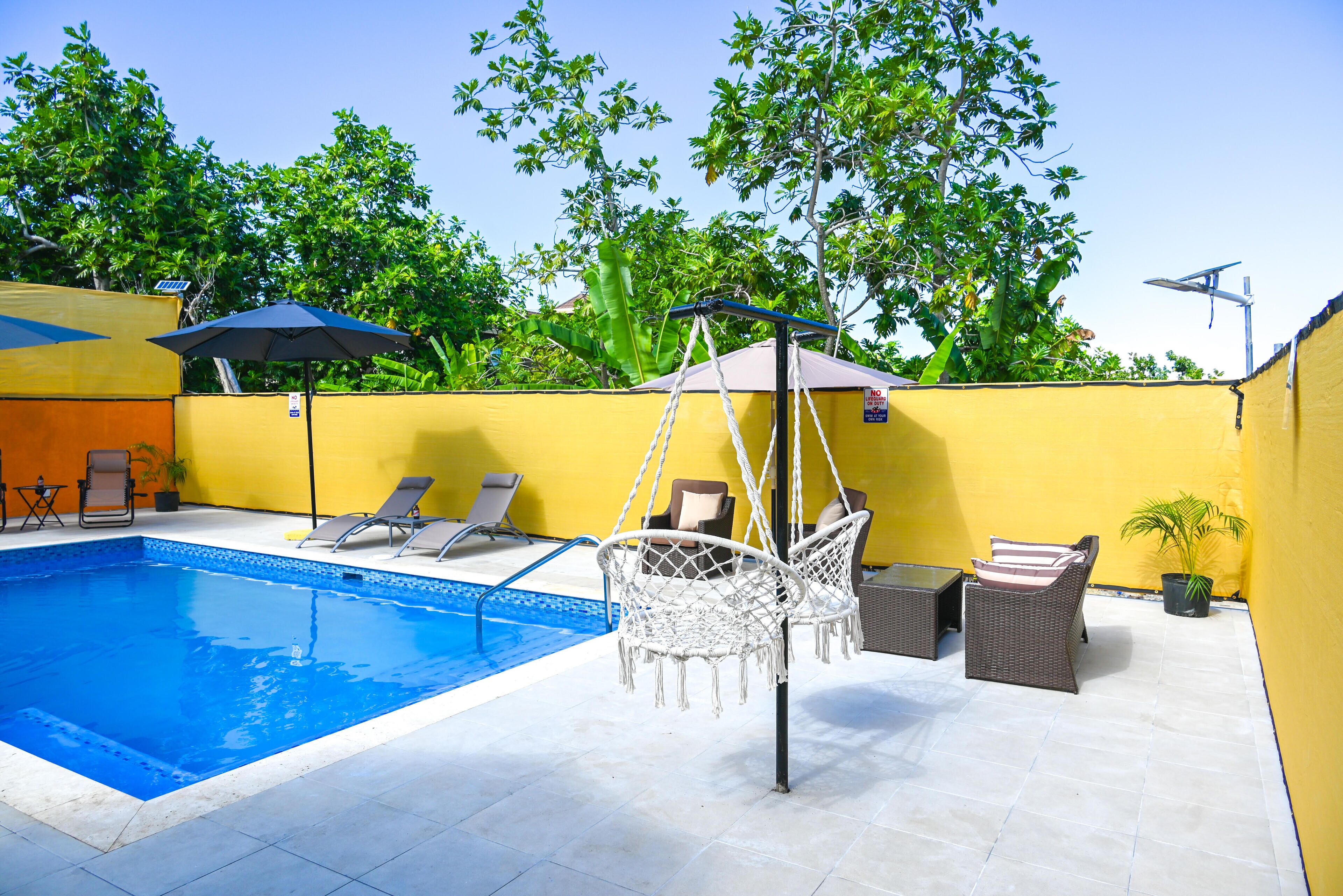 A swimming pool with lounge chairs and a hanging chair, surrounded by yellow walls and greenery.
