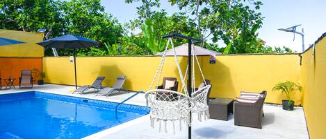 A swimming pool with lounge chairs and a hanging chair, surrounded by yellow walls and greenery.