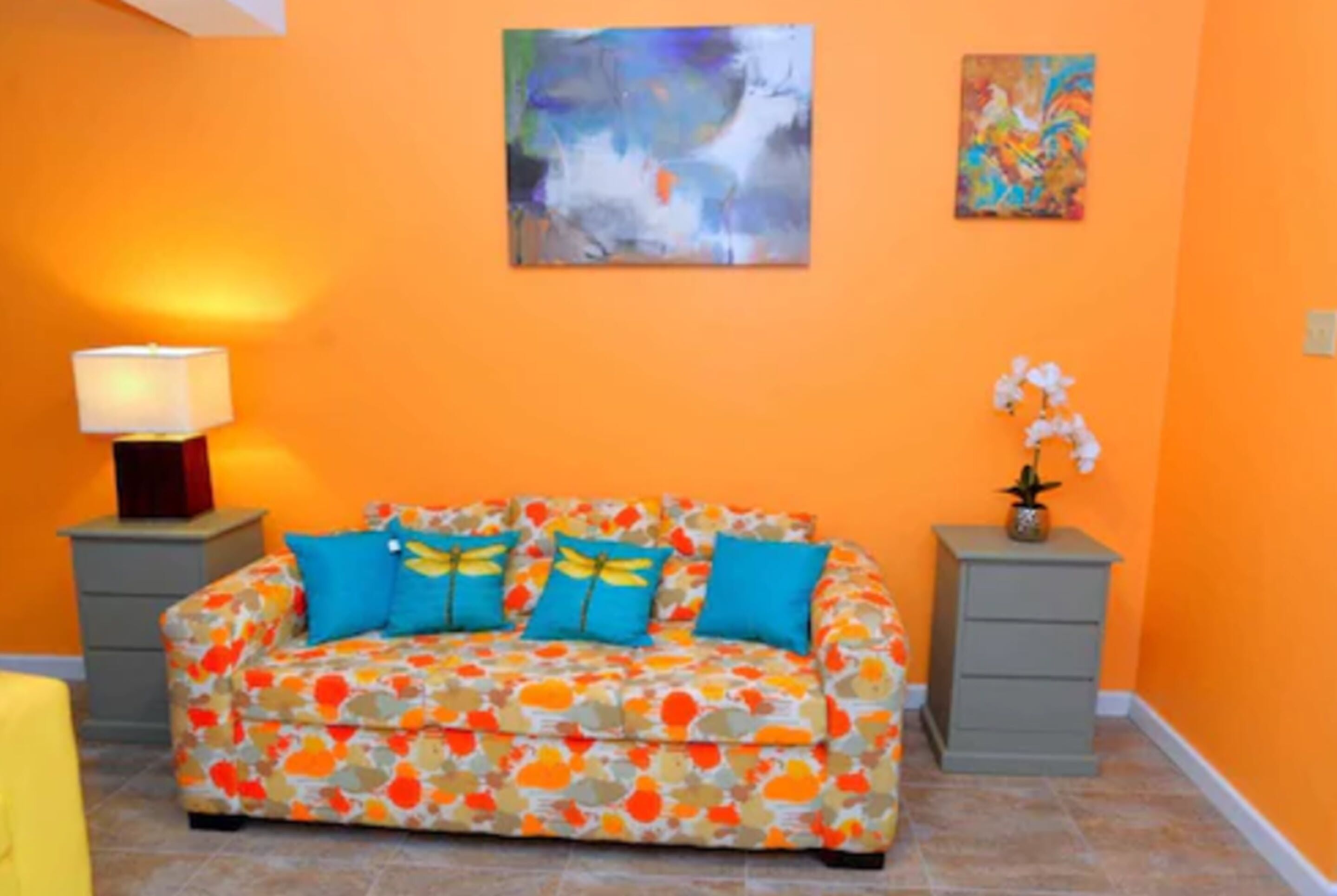 Une pièce avec un mur orange, un canapé agrémenté de coussins colorés, une lampe et une petite plante.