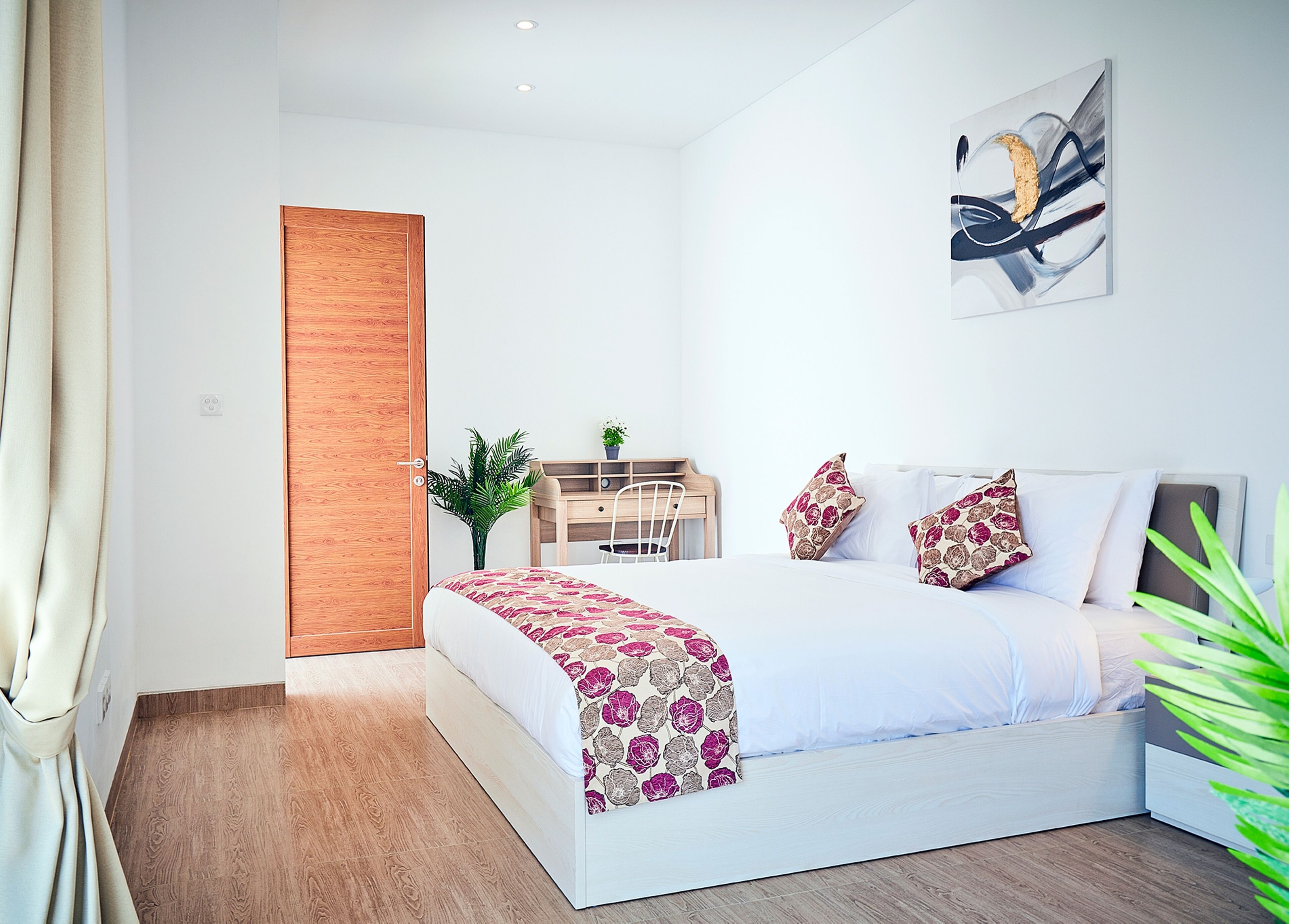 A modern bedroom with a wooden door, a desk, a chair, a bed with floral bedding, and a wall-mounted abstract painting.