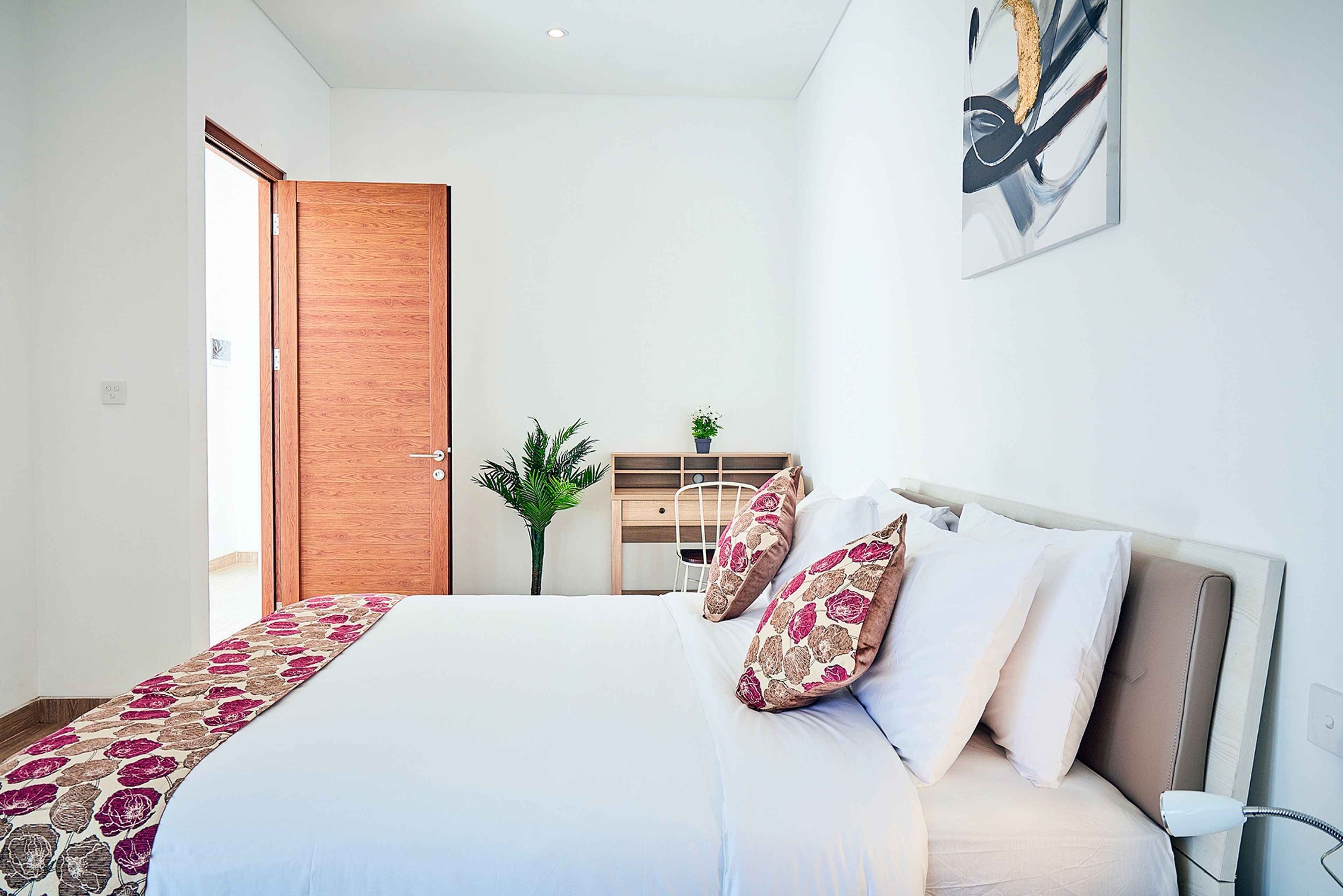 Un dormitorio ordenado con cama, almohadas, una mesita de noche, una planta y una puerta de madera.