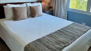 Een netjes opgemaakt bed met een witte quilt, een geweven plaid en een schilderij van boten aan de muur.