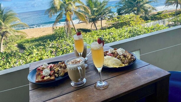 Een tafel met ontbijtproducten en drankjes op een balkon met uitzicht op een strand met palmbomen en de oceaan.