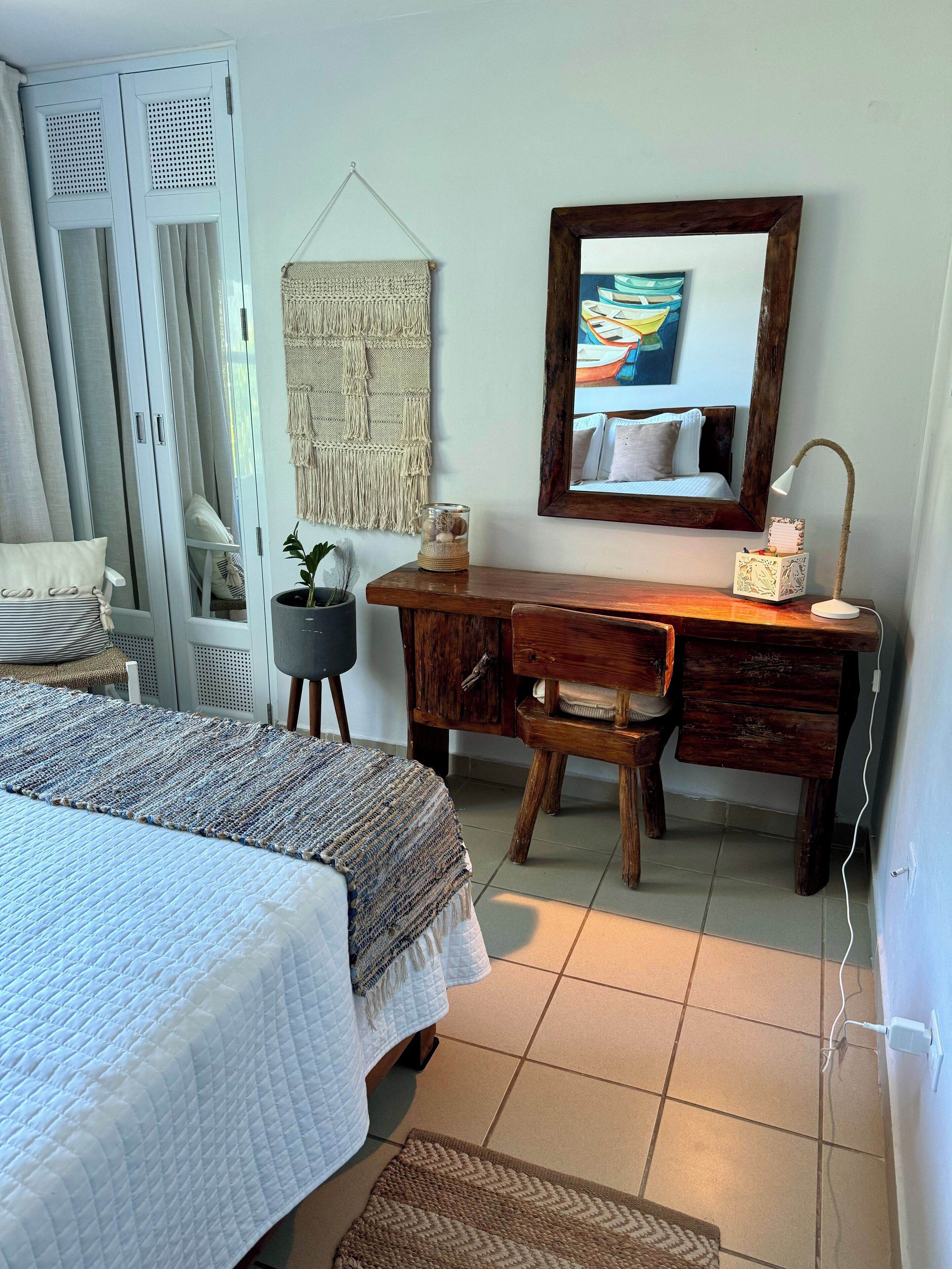 Un dormitorio con cama, escritorio, silla, un tapiz de macramé en la pared, un cuadro enmarcado y una lámpara de noche.