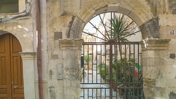 An arched stone doorway with a metal gate, a wooden door to the left, and a potted plant inside. - Dione monolocale quiet and confortable (Syracuse)