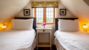 A bedroom with two beds, a nightstand with a vase of flowers, and a window with a view of greenery. - Kingfisher Cottage - sleeps 10 guests  in 5 bedrooms (Winchcombe)