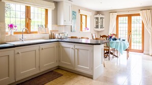 A kitchen with a central island, a dining area with a table, and a view of the outdoors through large windows. - Kingfisher Cottage - sleeps 10 guests  in 5 bedrooms (Winchcombe)