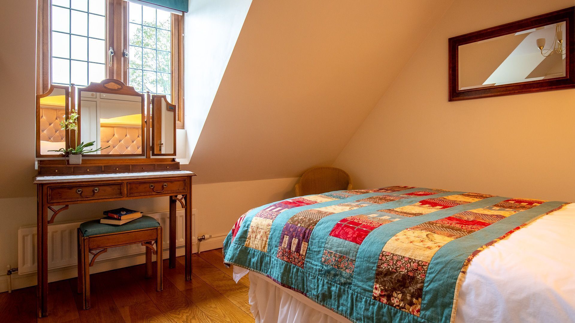 Un dormitorio con un escritorio de madera, una silla, una cama con colcha, un espejo y una ventana con vista a árboles.