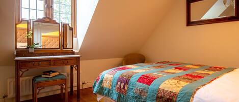 Un dormitorio con un escritorio de madera, una silla, una cama con colcha, un espejo y una ventana con vista a árboles.