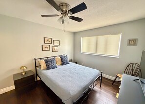 Une chambre à coucher avec un lit, un ventilateur de plafond, une fenêtre avec des stores et des tableaux encadrés accrochés au mur.
