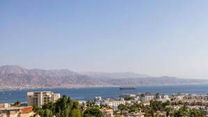 A coastal cityscape with a large ship in the harbor, buildings, and mountains in the background. - YalaRent Sunora 3BR APT - Families only (Eilat)