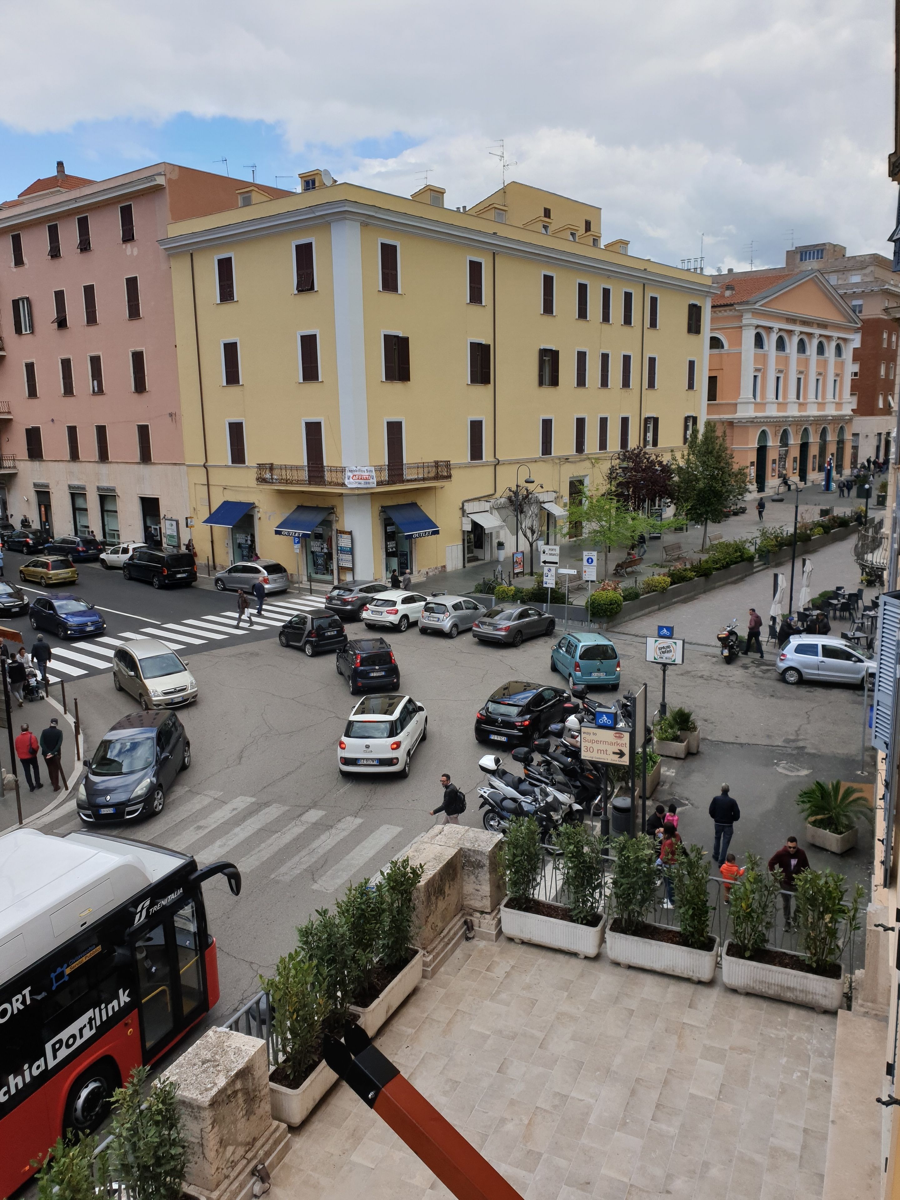 Una calle urbana concurrida, con coches aparcados y un autobús. Se ven edificios de varios pisos y balcones.