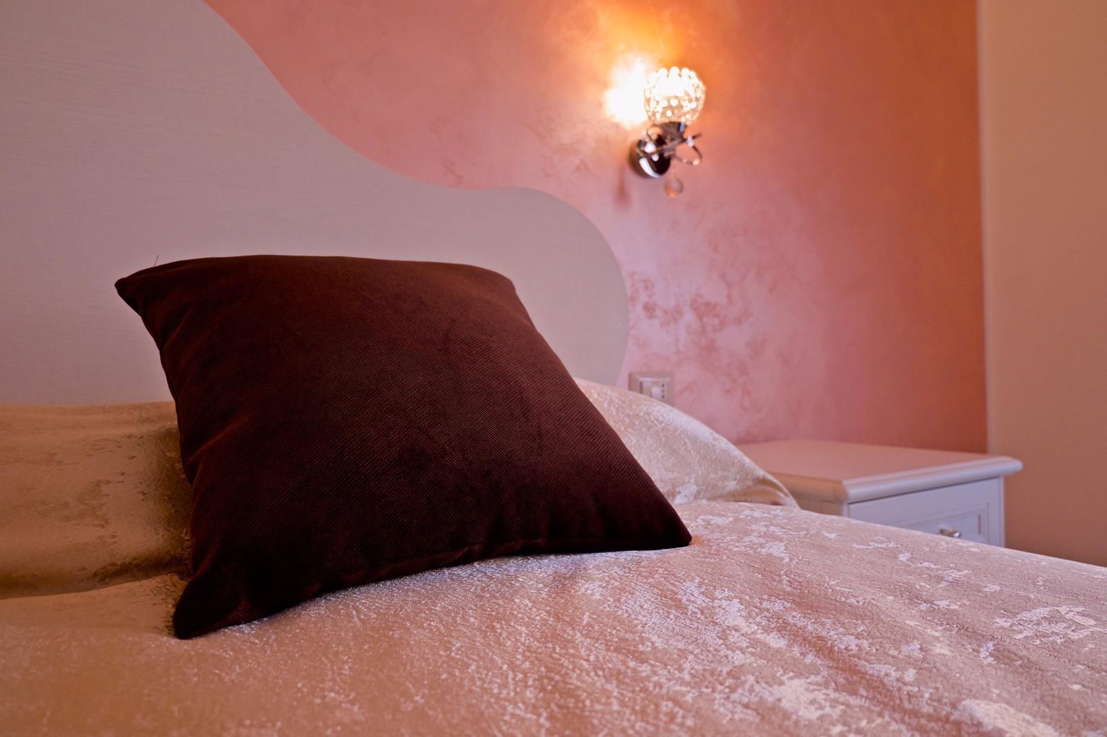 Ein Bett mit einem dunkelbraunen Kopfkissen und einer weißen Bettdecke in einem Zimmer mit rosafarbener Wand und einer Wandleuchte.