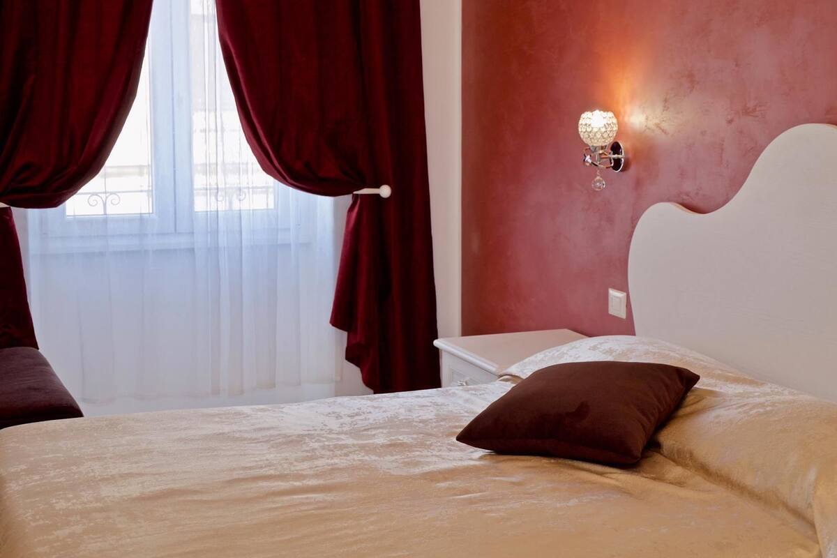 A hotel room with a bed, a nightstand, a wall-mounted lamp, and a window with sheer curtains.