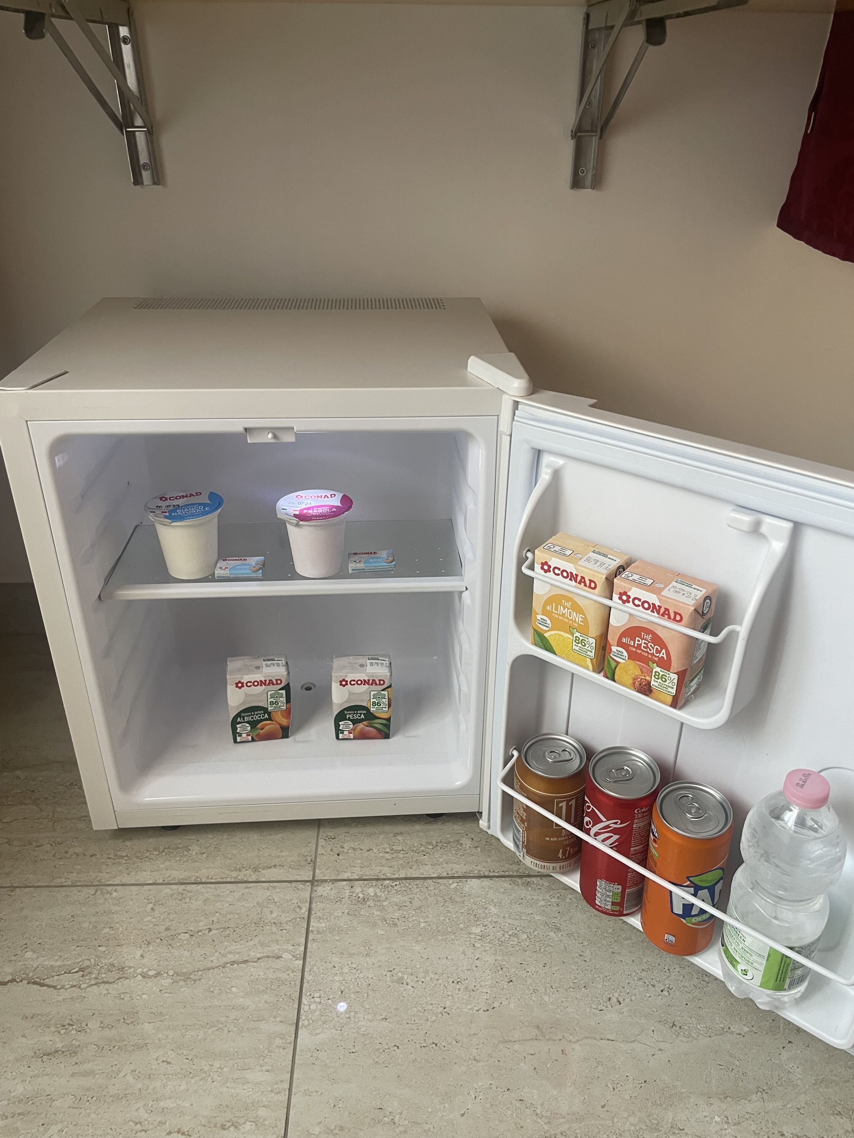 Geladeira aberta com prateleiras contendo caixas de suco Conad, latas de Coca-Cola e uma garrafa de água.