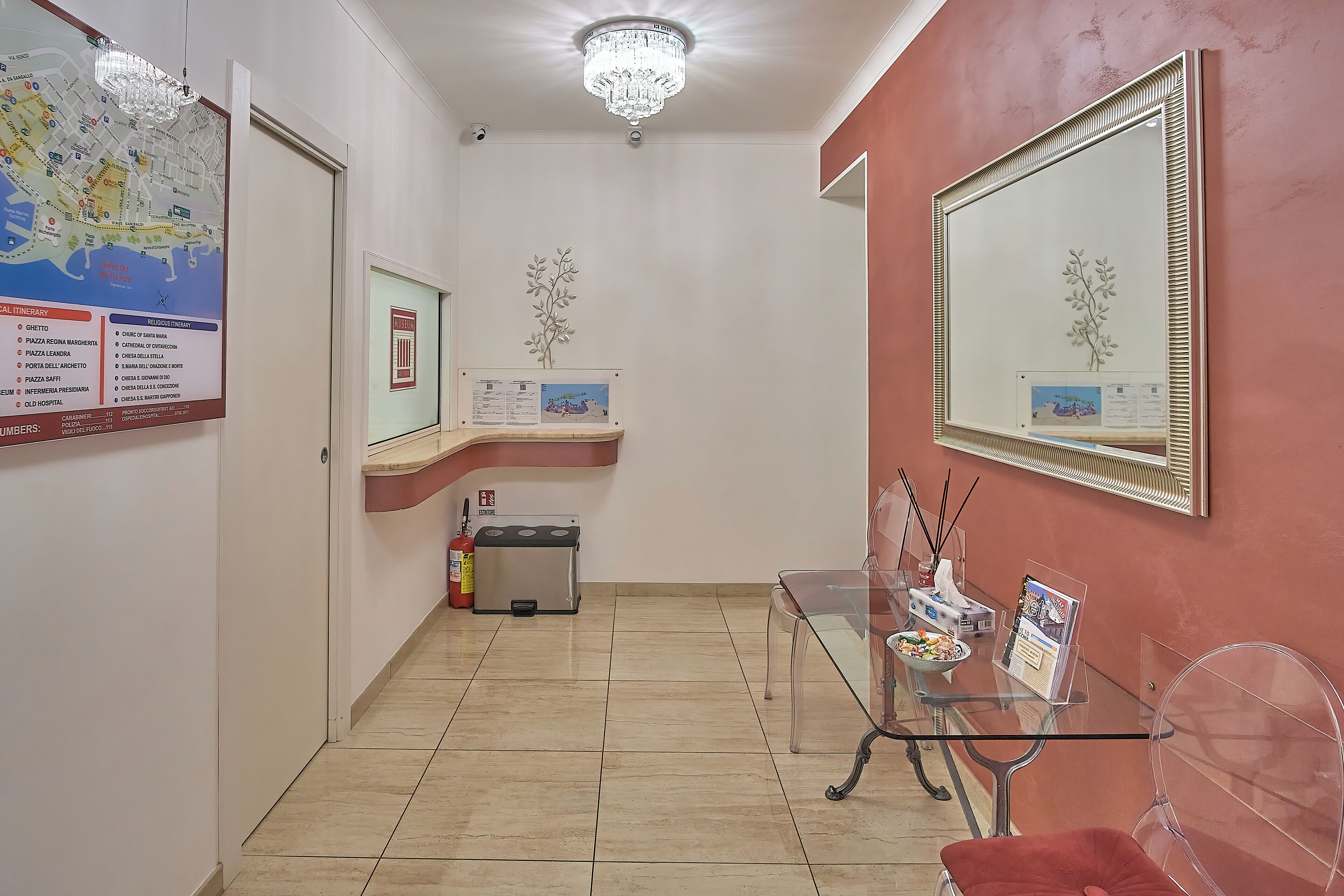 Pasillo de hotel con un mapa, una araña, un espejo, extintor y una mesa de vidrio con sillas.