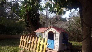 A small playhouse with a red roof and blue door, surrounded by a wooden fence, set in a grassy area with trees in the background. - My holiday refuge in the Northeast of Mallorca (Font de Sa Cala)