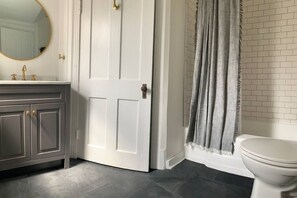 Ein Badezimmer mit einer weißen Tür, einem grauen Waschtisch mit goldenen Griffen, einem runden Spiegel und einer weißen Toilette.