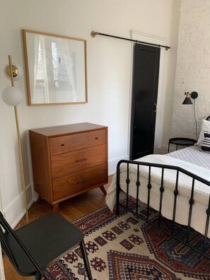 Ein Schlafzimmer mit einem Bett, einer Holzkommode, einem Sessel, einem gemusterten Teppich, einer Wandlampe und einer Tür.