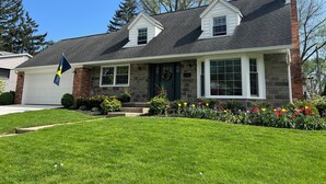 A two-story house with a stone facade, a front porch, and a flag. - Fantastic 4-Bedroom Family Home - Short Walk to Michigan Stadium (Ann Arbor)