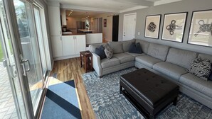 A modern living room with a grey sectional sofa, a black ottoman, and a patterned rug. A glass door leads to a kitchen with white cabinets and a stainless steel refrigerator. - Fantastic 4-Bedroom Family Home - Short Walk to Michigan Stadium (Ann Arbor)