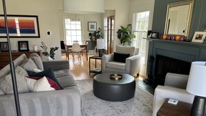 A modern living room with a grey sofa, a round black ottoman, and a patterned rug. - Fantastic 4-Bedroom Family Home - Short Walk to Michigan Stadium (Ann Arbor)
