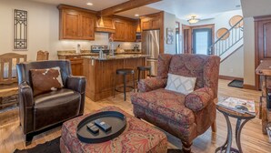 A spacious living room with a large leather armchair, a patterned sofa, and a coffee table with remotes. A kitchen with wooden cabinets and a stainless steel refrigerator is visible in the background.