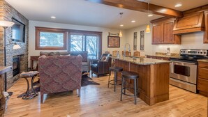 A spacious kitchen with wooden cabinets, a large island with stools, a stone fireplace, and a cozy seating area with patterned upholstery.