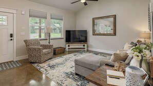 A living room with a ceiling fan, a television on a stand, a patterned armchair, a grey ottoman, a coffee table with a notebook, a vase with flowers, and a large mirror on the wall.
