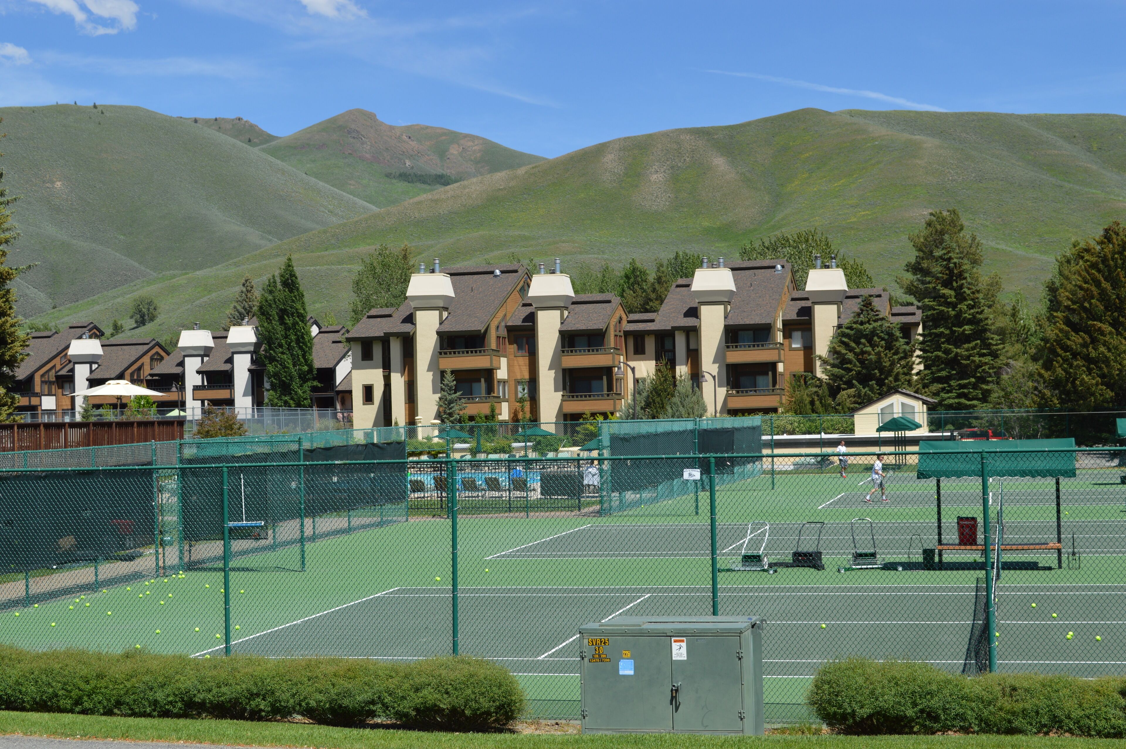 Una cancha de tenis rodeada de una valla, con montañas al fondo y edificios residenciales.