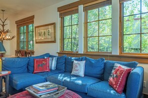 Ein gemütliches Wohnzimmer mit einem blauen Sofa, einem Kamin und großen Fenstern mit Blick auf Grünflächen.