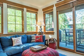 Una sala acogedora con un sofá azul, un reposapiés rojo y una lámpara. Hay ventanas grandes con marcos de madera y vistas al exterior lleno de vegetación.