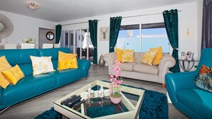 Un salon moderne avec des canapés de couleur turquoise, une table basse en verre et une vue sur la mer grâce à de grandes fenêtres.