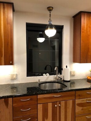 Una cocina con armarios de madera, encimera negra, un lavamanos y una lámpara colgante.