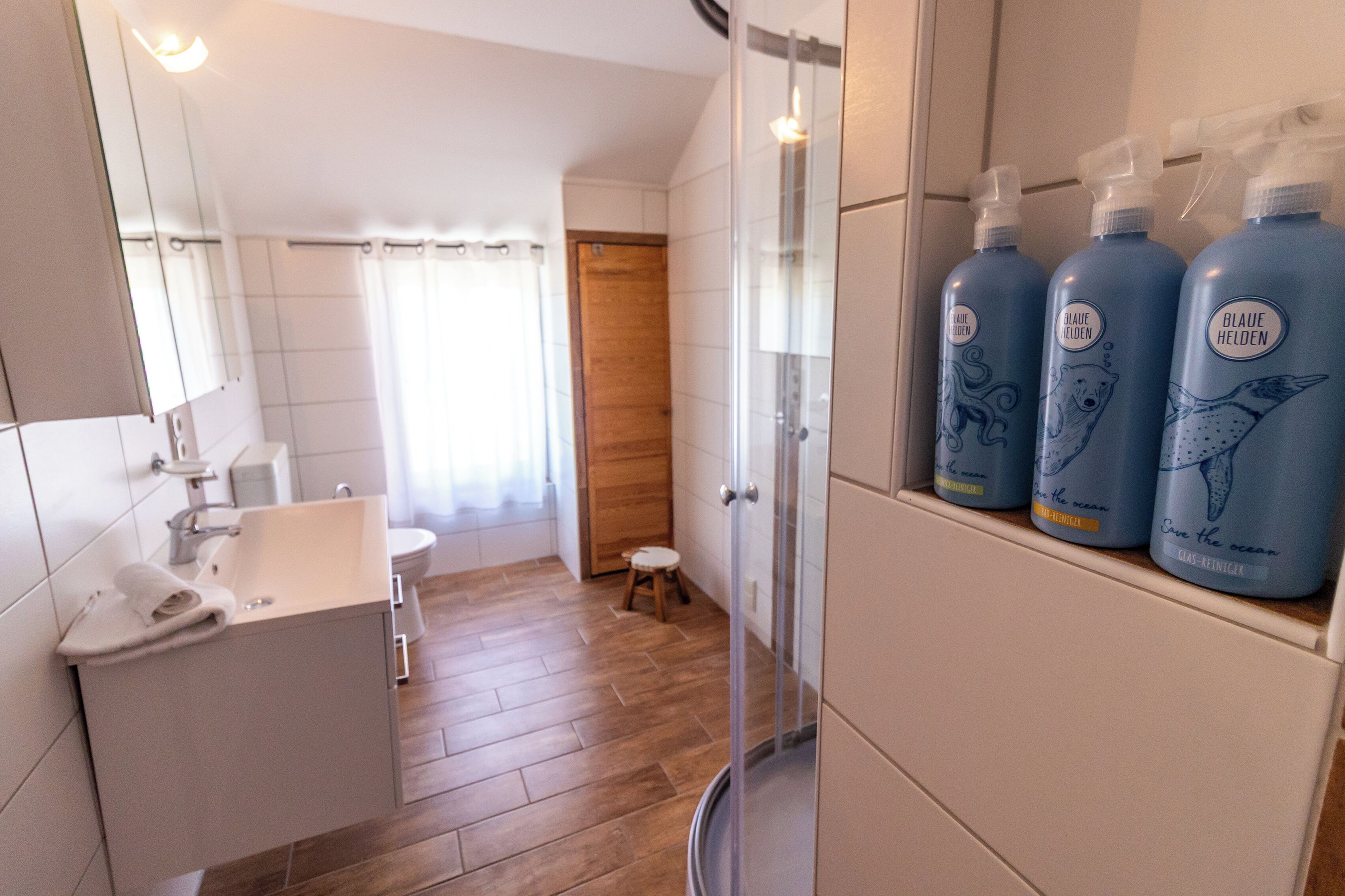 A bathroom with a wooden floor, a sink, a toilet, and a shower. There are three bottles of 'Blaue Helden' on a shelf.